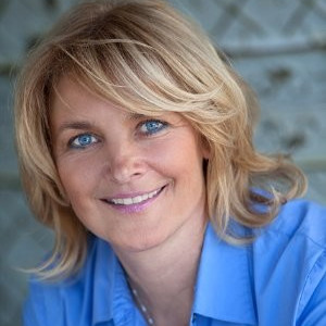 A headshot of Denise Clements, a white woman with bright blue eyes and shoulder-length blonde hair. She wears a blue shirt with slight purple undertones that bring out the color in her eyes. She also wears a thin necklace. It looks like it has a pendant, but it is out of frame.