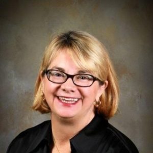 A headshot of Shari Frazier, a white woman with chin-length blonde hair. She wears rectangular glasses with black frames. She wears a black collared shirt.