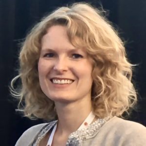 A headshot of Katrina Mobius, a white woman with blonde curly hair that just brushes the top of her shoulders. She wears an off-white cardigan and has a white lanyard around her neck.