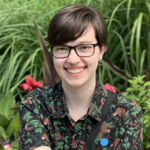 A headshot of Sam Mankowski, a white person with short brown hair. They have brown eyes behind big rectangular glasses with black frames. They are wearing a button-down shirt with sloths on it. They are sitting in front of lush green plants. Some pink flowers are visible over their shoulders.